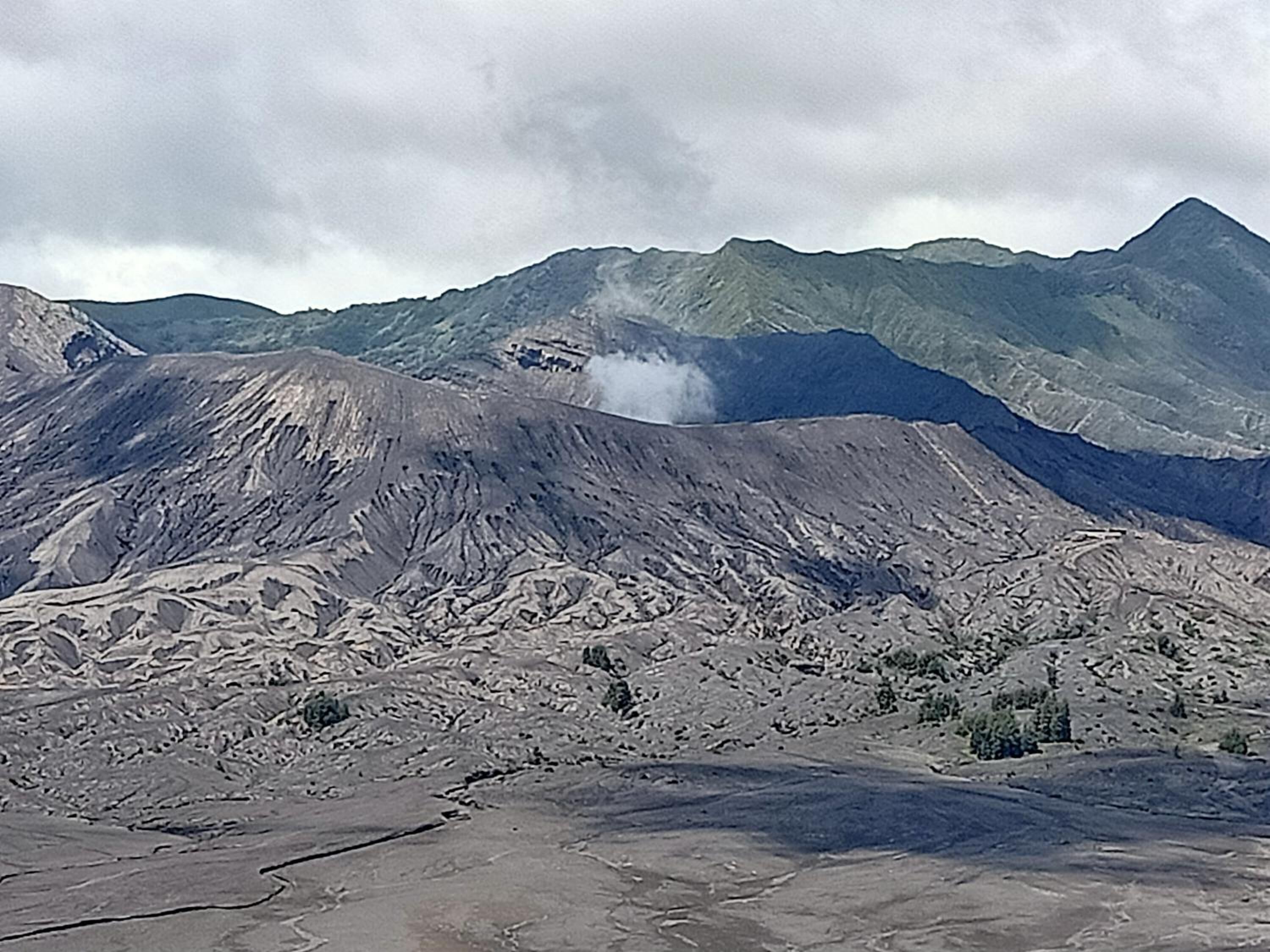Un paysage montagneux accidenté, dominé par un sommet imposant d’où s’échappe de la fumée, entouré d’un terrain aride et de collines verdoyantes à l’horizon.