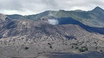 Un paysage montagneux accidenté, dominé par un sommet imposant d’où s’échappe de la fumée, entouré d’un terrain aride et de collines verdoyantes à l’horizon.