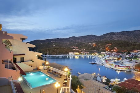 A hotel with a swimming pool, outdoor seating, and a view of a harbor with boats and a mountain backdrop.