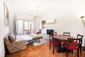 A compact room with a bed, a dining table with chairs, a TV, and a chandelier.