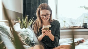 Una persona sentada en una cama en una habitación con ventana, sosteniendo un teléfono inteligente.