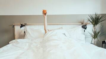 Una cama con sábanas blancas, dos lámparas de noche y una planta en maceta a la derecha.