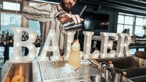 Un bartender preparando una bebida en un bar, con letras iluminadas que deletrean "BAILLER" al fondo.