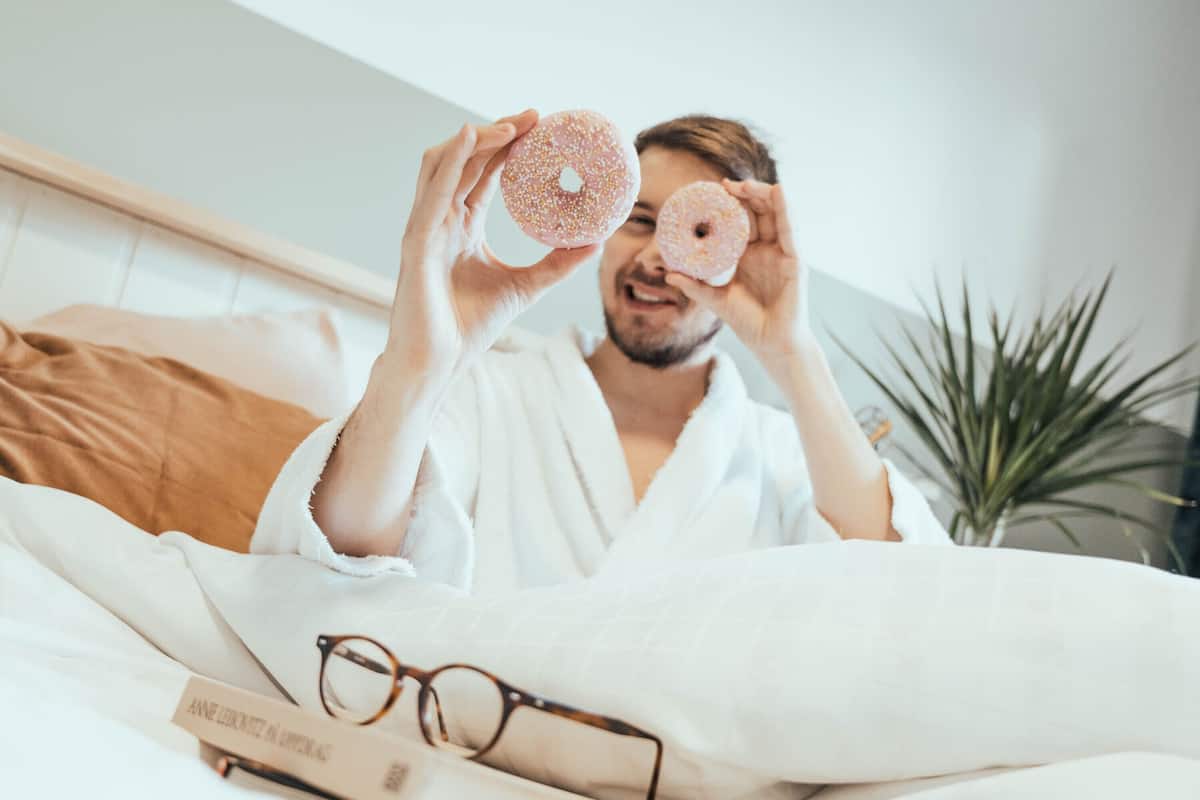 A person in a white robe holding two donuts, with a book and glasses on a bed.