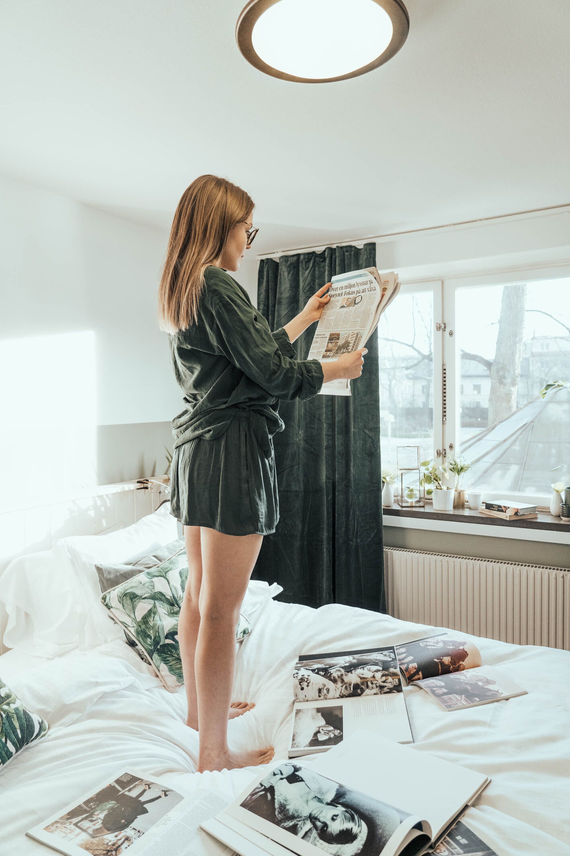 A person standing on a bed reading a book in a room with a window, a radiator, and a curtain.