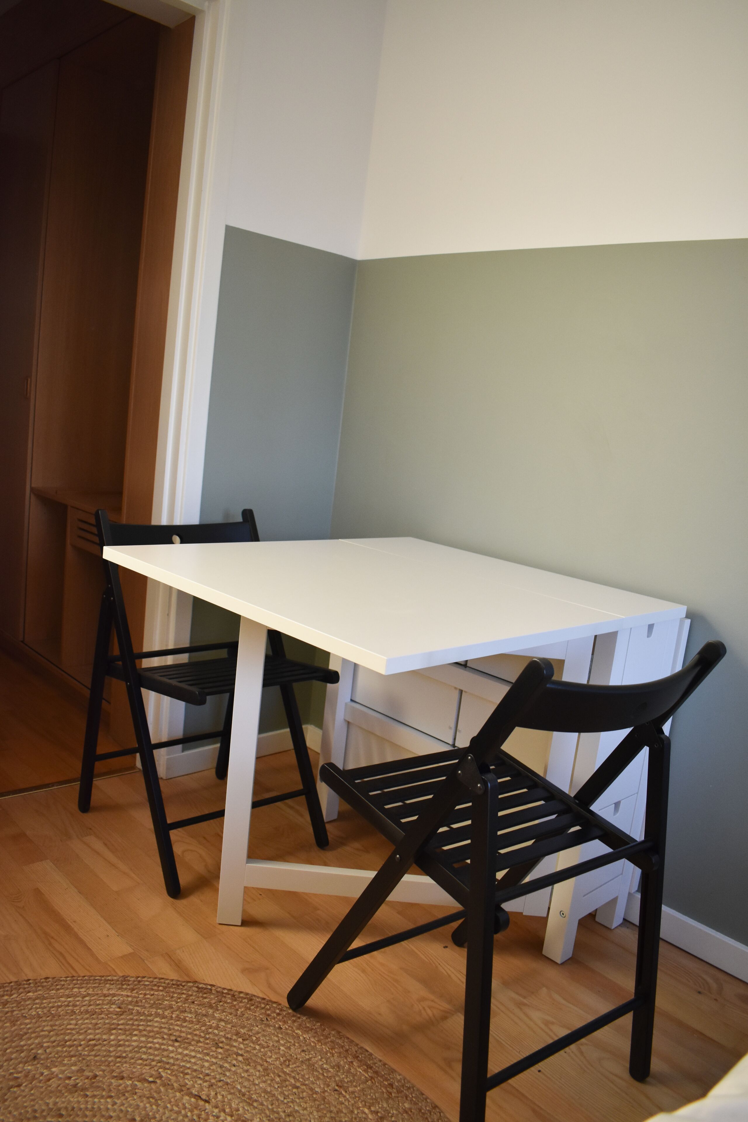 Una mesa blanca con dos sillas negras en una habitación con piso de madera y paredes de color claro.
