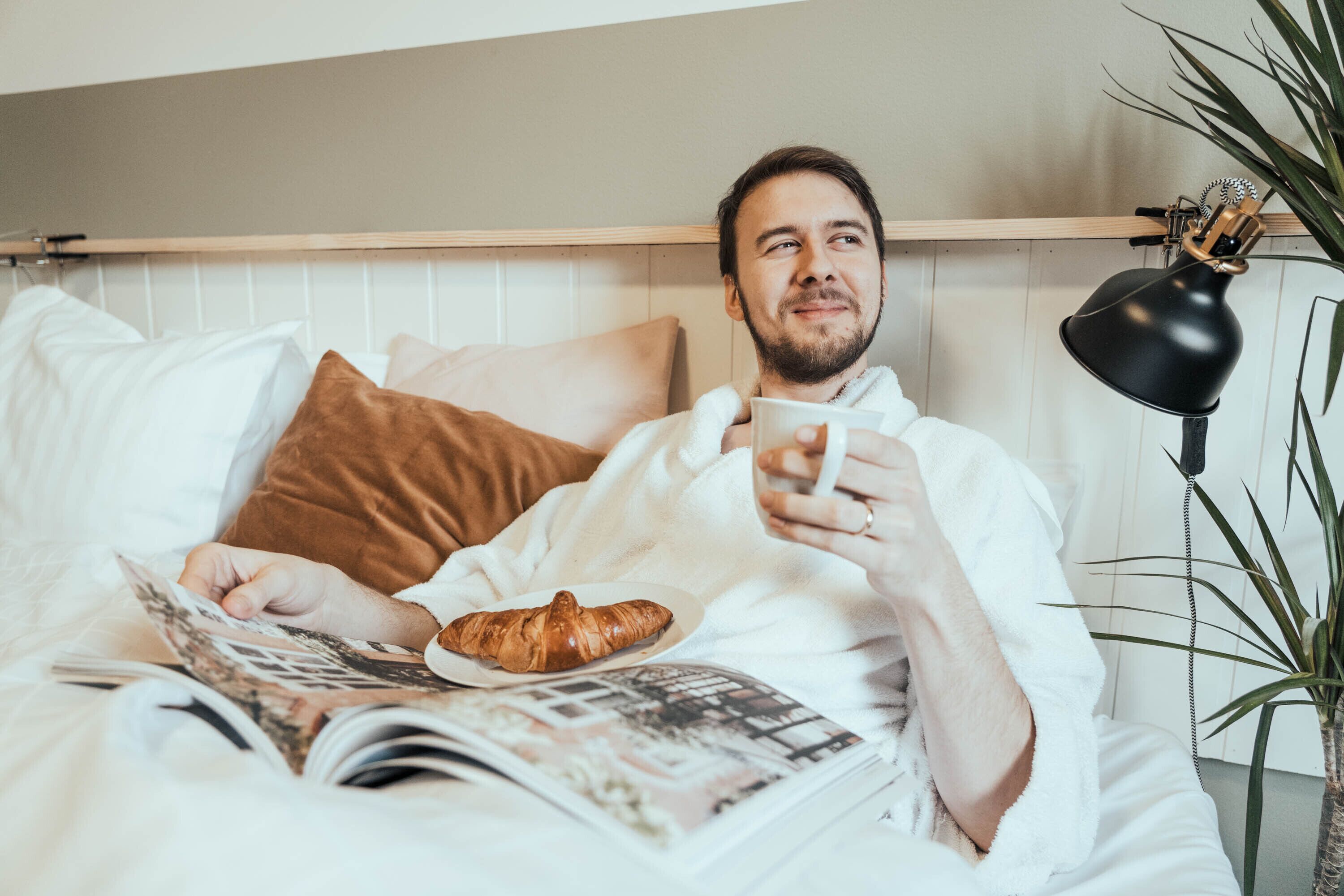 A person lying in bed with a book and a croissant, a lamp, and a plant in the room.