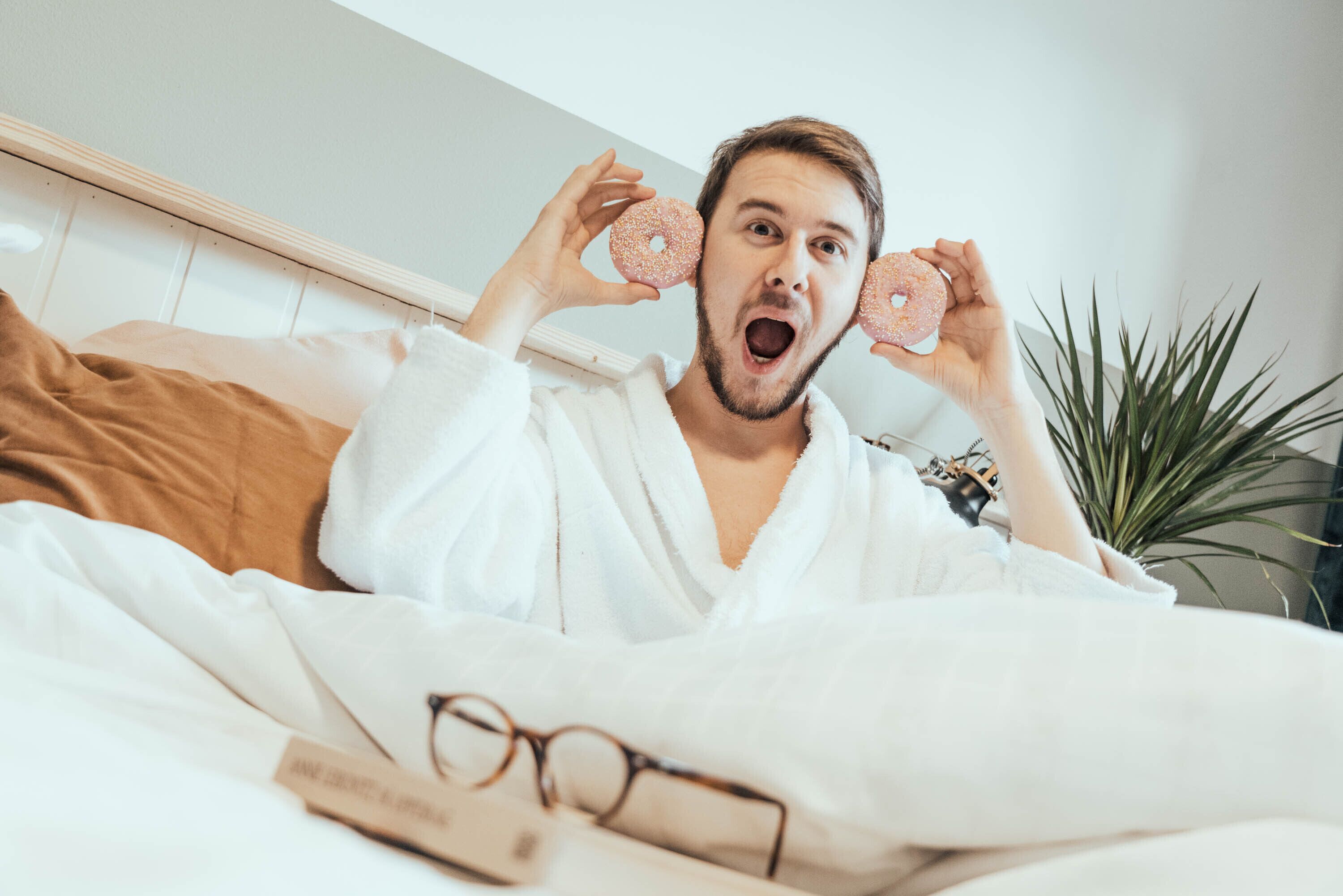 A person in a white bathrobe lying in bed with a plant and a pair of glasses on the bed.