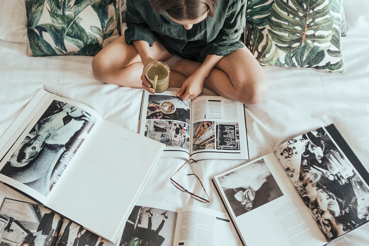 A person sitting on a bed with a book and a cup, surrounded by magazines and a pair of glasses.