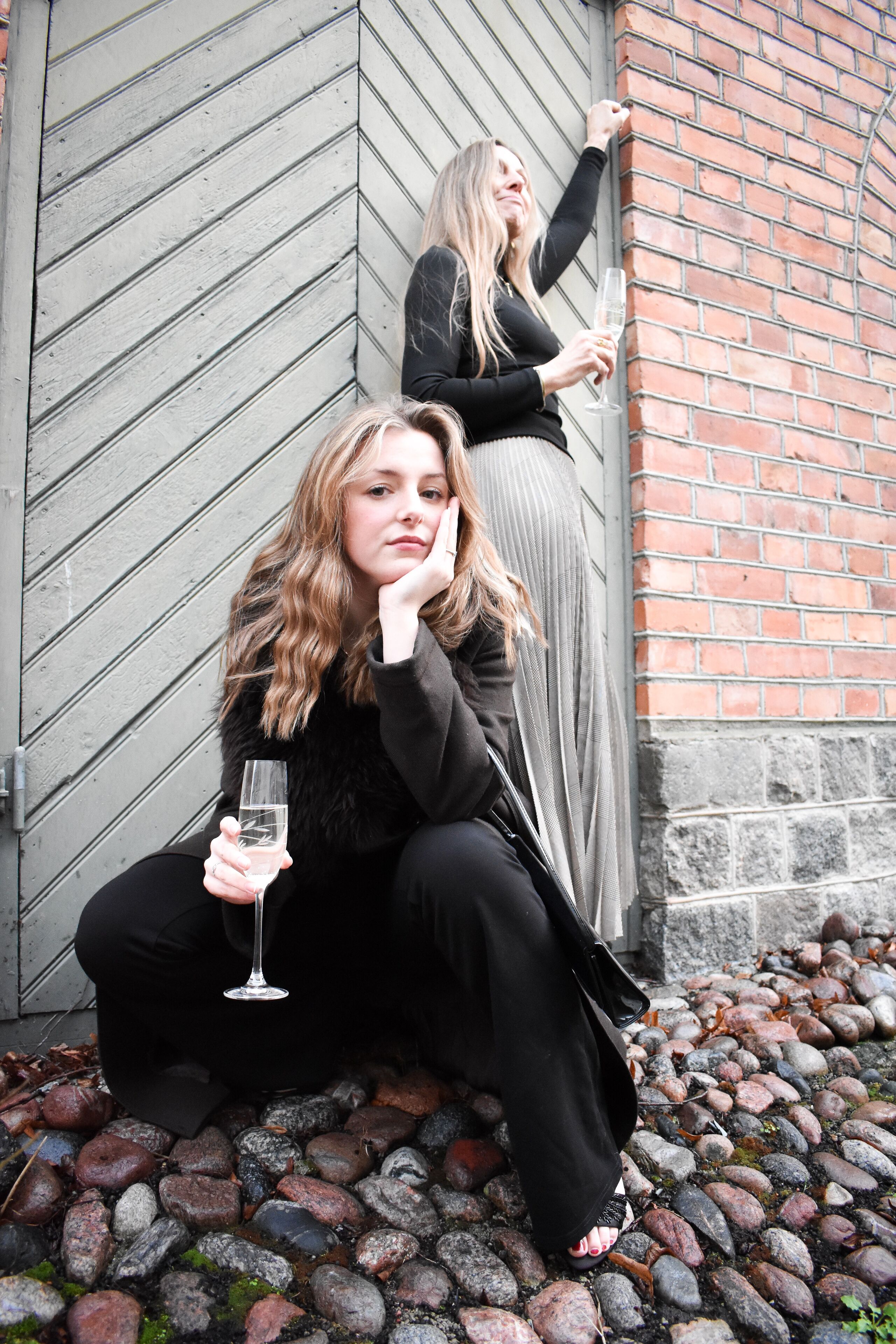 Two individuals sitting on a stone-paved area with a brick wall and a wooden door in the background.