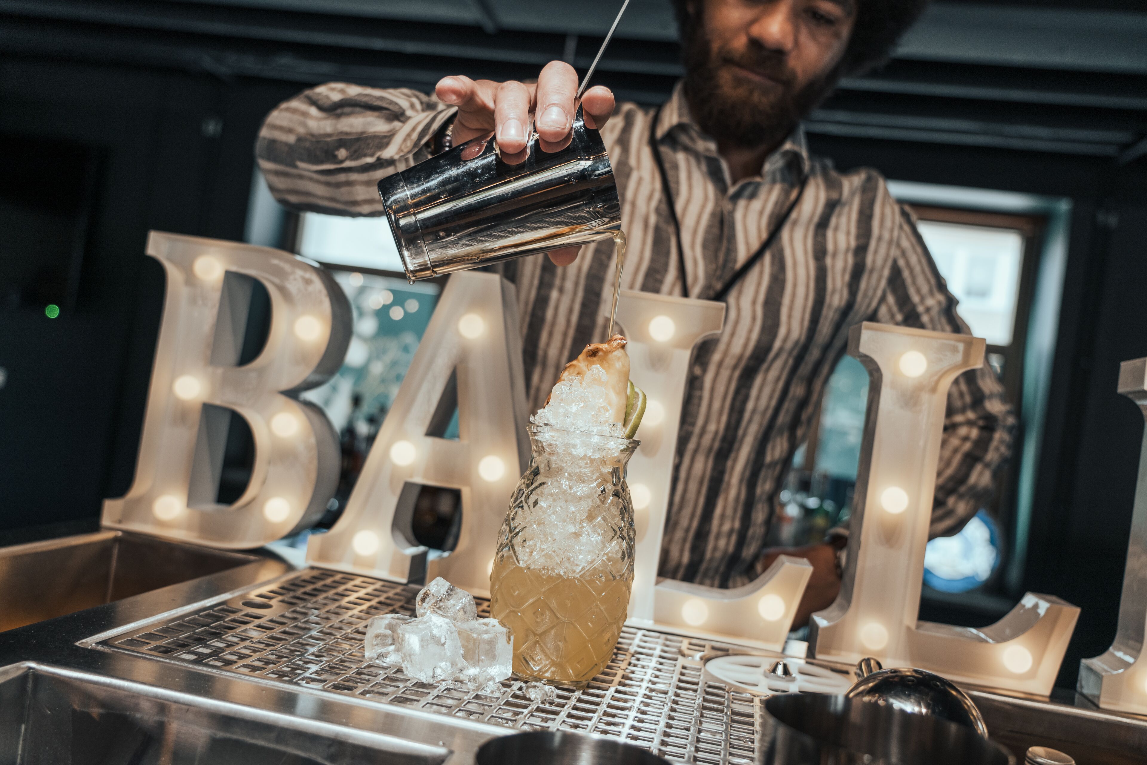 Ein Barkeeper gießt ein Getränk in ein Glas mit Eiswürfeln. Im Hintergrund leuchten die Buchstaben 'BALL'.