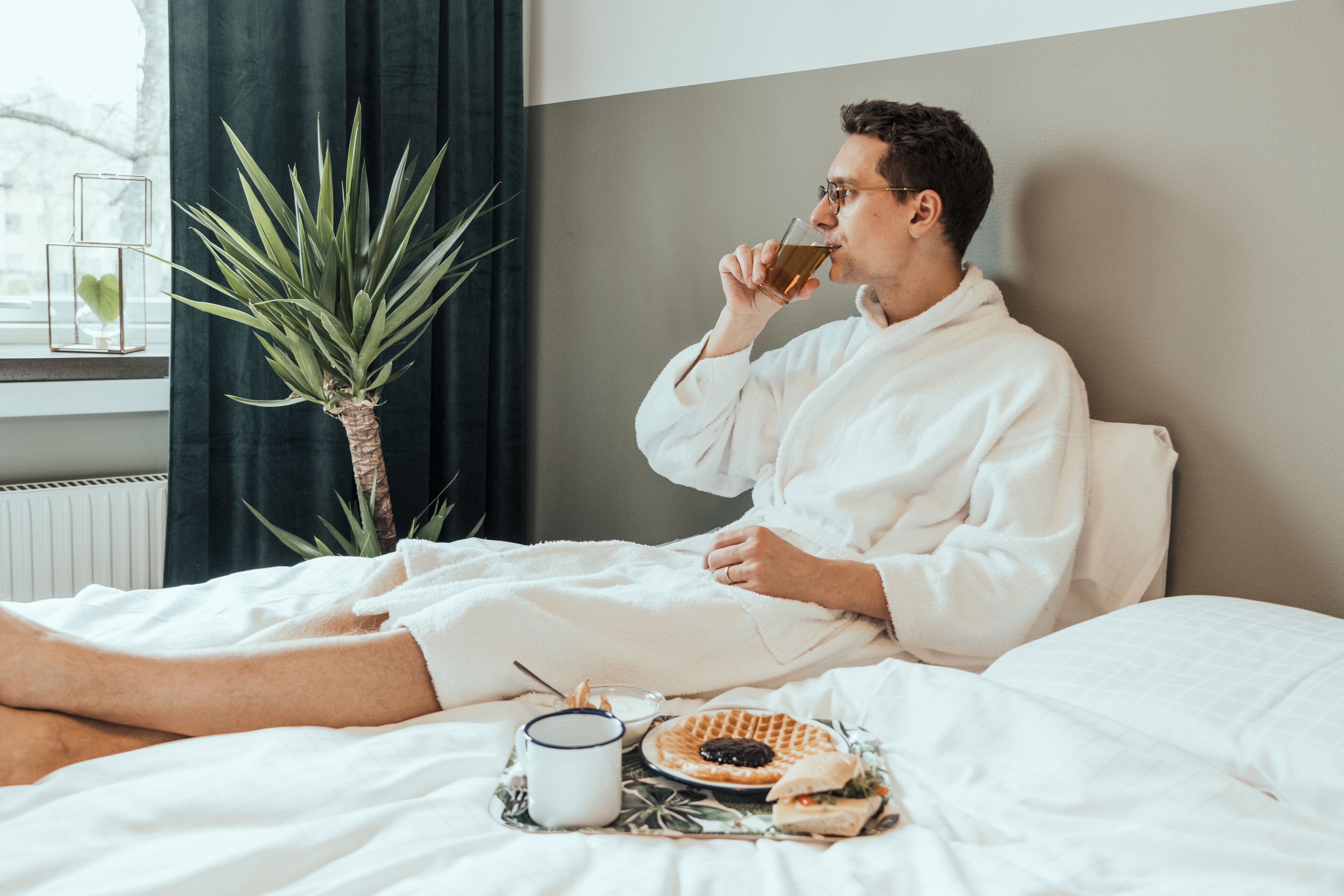 A person in a white bathrobe sitting on a bed with a tray of food and a cup, near a window with a plant.