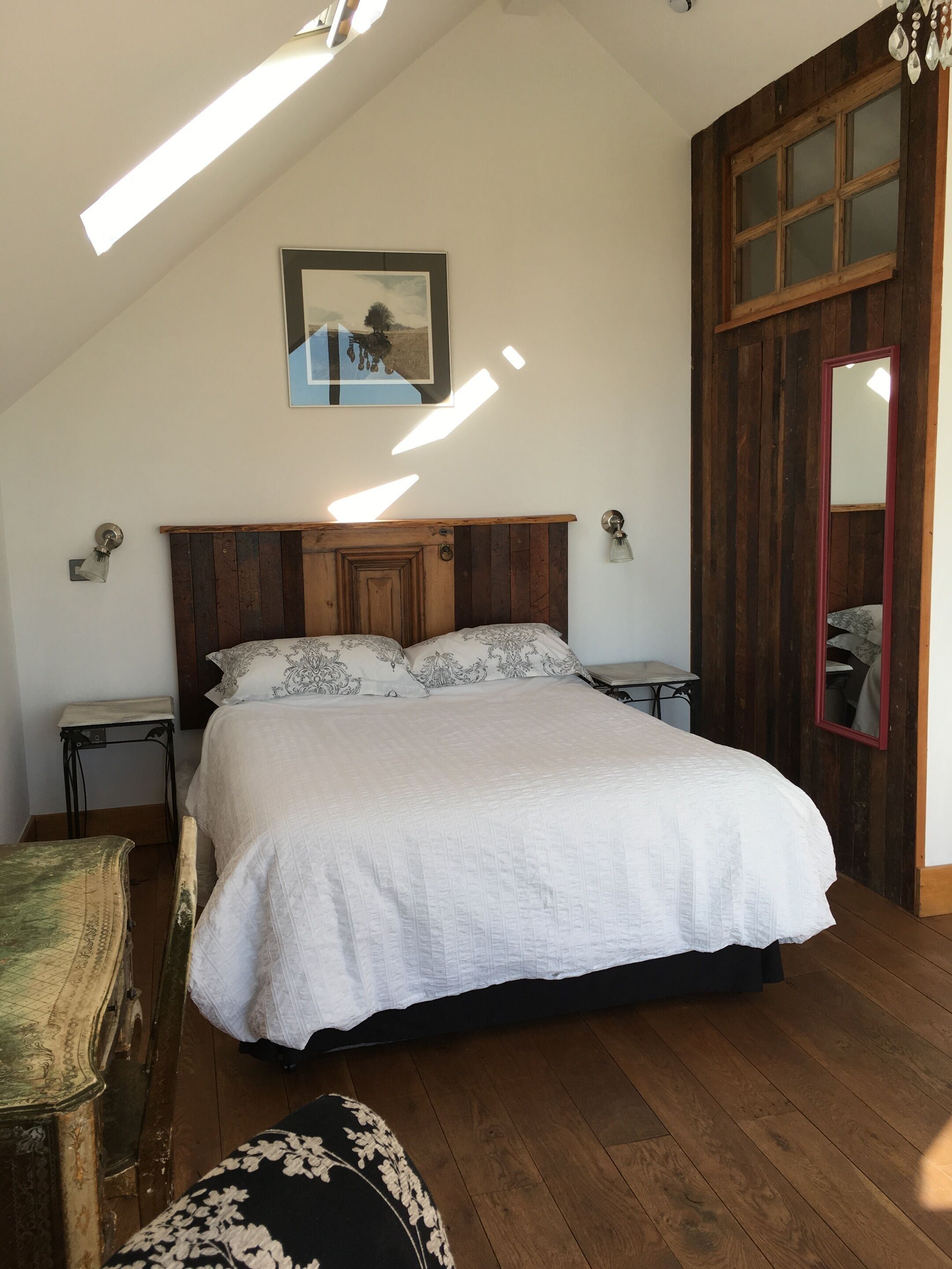 A bedroom with a wooden headboard, a bed with white bedding, a small table, a chair, and a mirror.