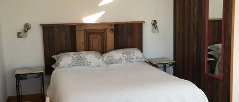 A bedroom with a wooden headboard, a bed with white bedding, a small table, a chair, and a mirror.