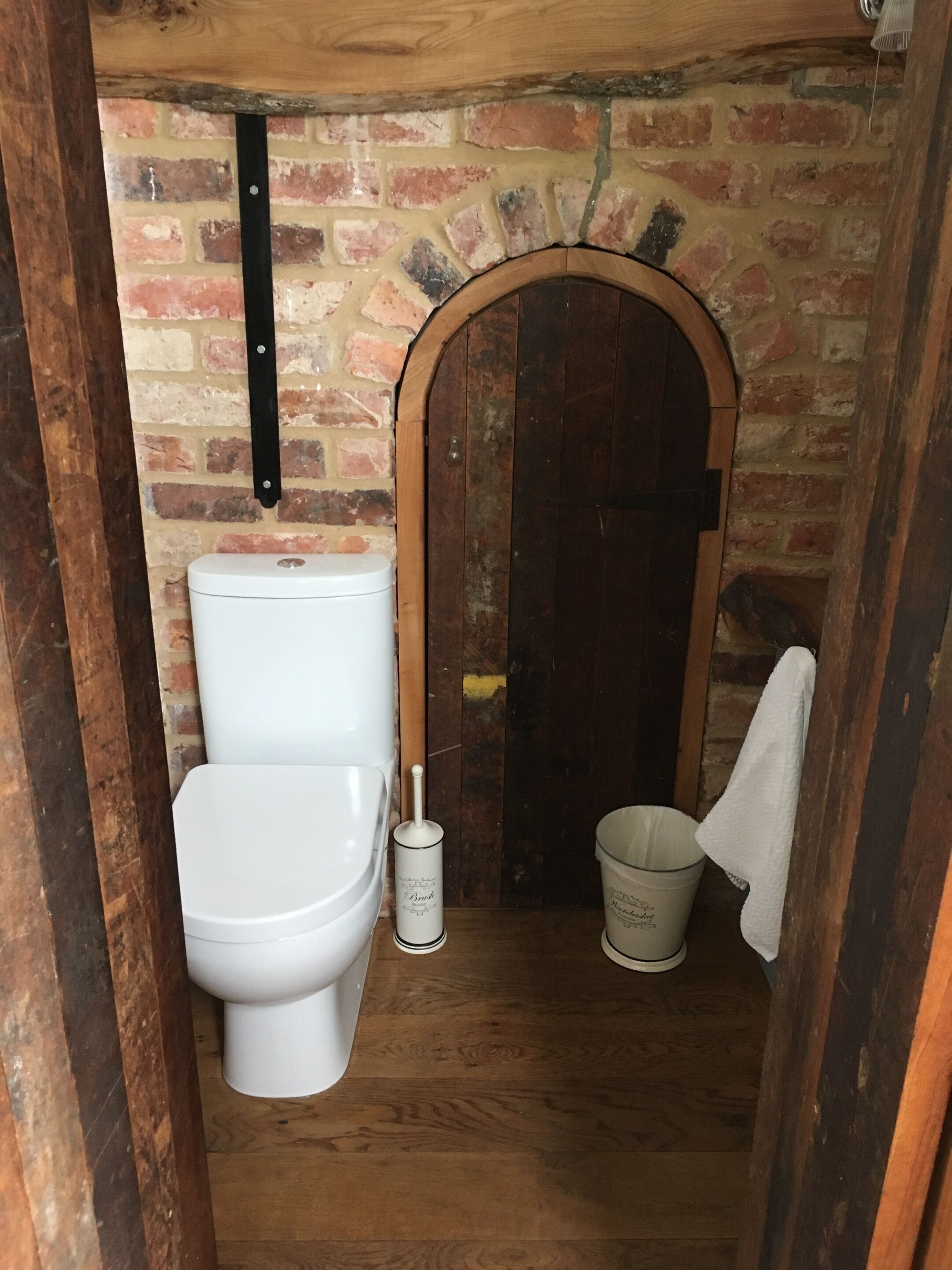 A small, rustic bathroom with a wooden door, a white toilet, and a white bucket.