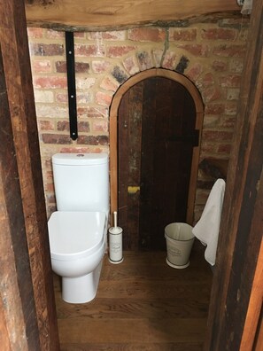 Un baño pequeño y rústico con una puerta de madera, un inodoro blanco y un balde blanco.
