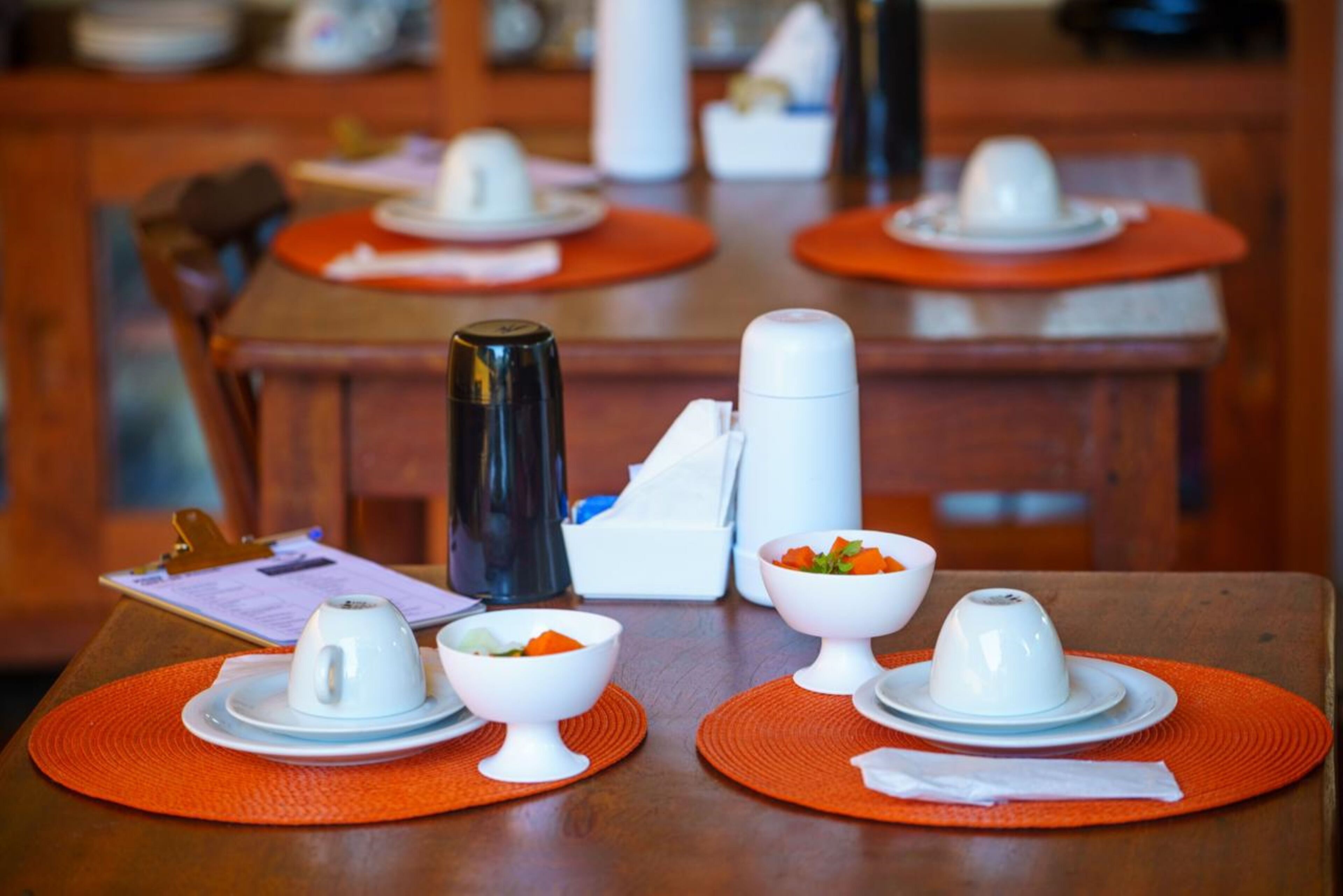 A dining table with white cups and bowls on orange placemats, a clipboard, and a thermos.