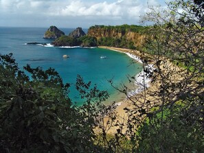 A coastal landscape with a beach, rocky cliffs, and boats on the water. - Pousada Guedes Noronha (Fernando de Noronha)