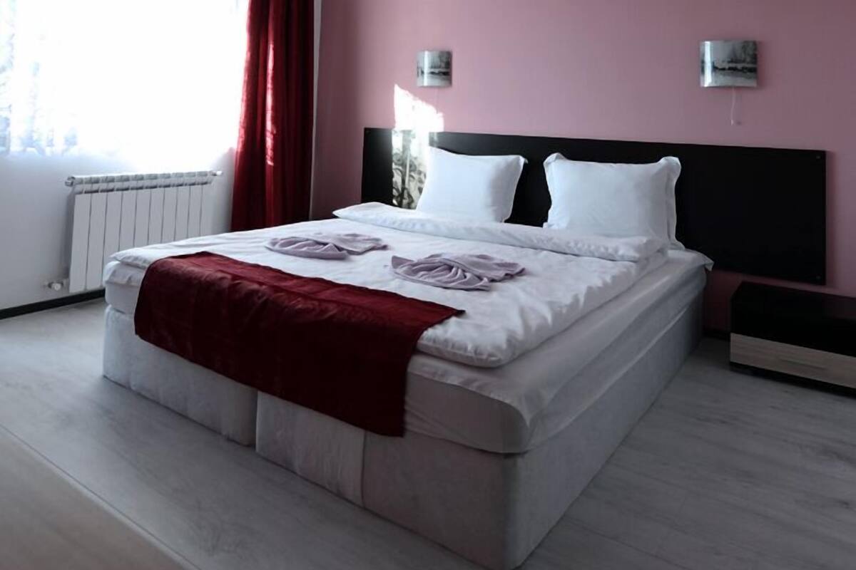 Una cama bien hecha con sábanas blancas y un plaid rojo, cabecera negra y dos apliques de luz en la pared.