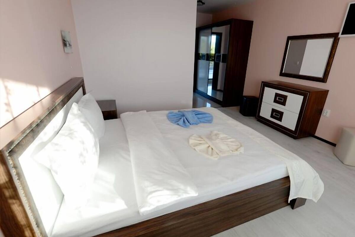 Una cama bien tendida con una toalla azul y una toalla blanca, un aparador de madera con cajones, un espejo y un armario en una habitación con paredes rosa claro.