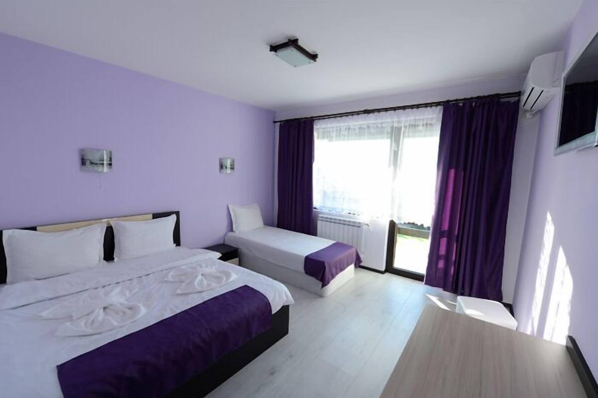 Habitación de hotel con dos camas, un escritorio de madera, una ventana con cortinas moradas y un equipo de aire acondicionado.