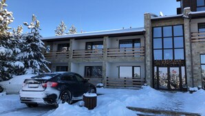 Un edificio de varios pisos con un balcón, un automóvil estacionado adelante y árboles cubiertos de nieve.