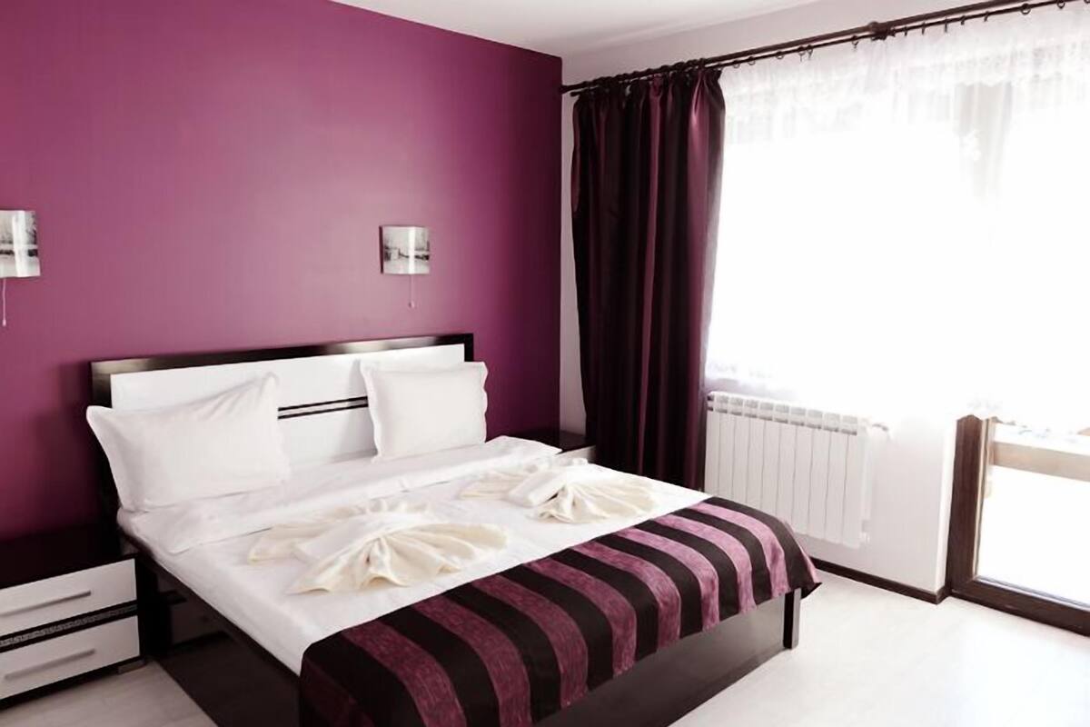 Habitación de hotel moderna con una cama grande, paredes moradas y vistas al exterior.