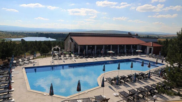 Una zona de piscina con sillones de descanso y sombrillas, un edificio con tejado rojo, y una vista de un lago y montañas al fondo.