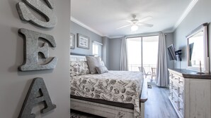 Een slaapkamer met een bed, een dressoir, een plafondventilator en een groot raam met gordijnen.