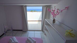Una habitación con balcón, cama con ropa de cama rosa, una silla, un jarrón con flores y una estantería con un lazo amarillo.