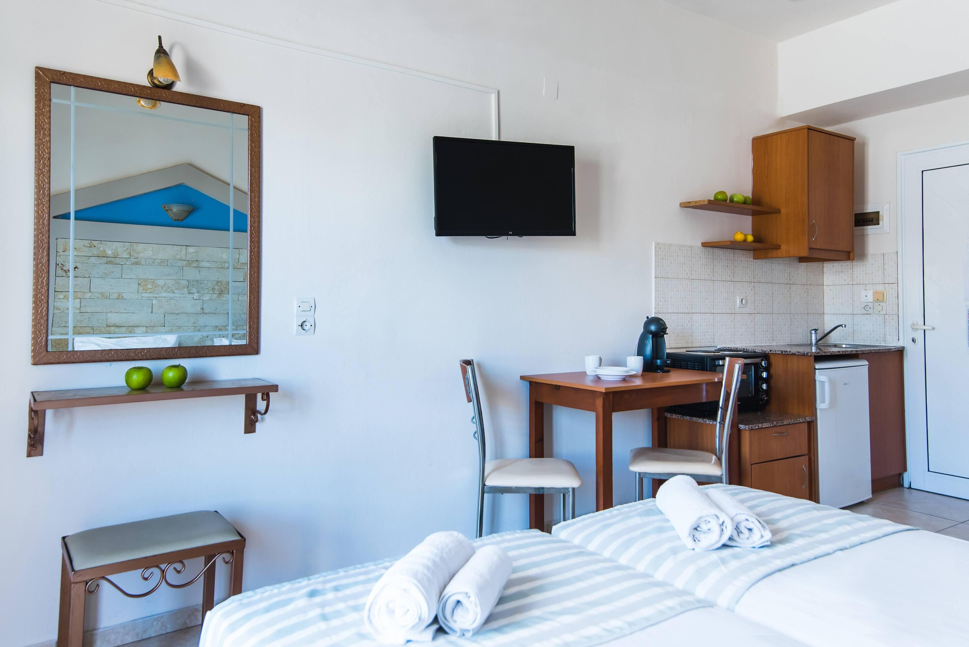 A hotel room with a bed, a desk with a chair, a small table, a television mounted on the wall, and a kitchenette area.