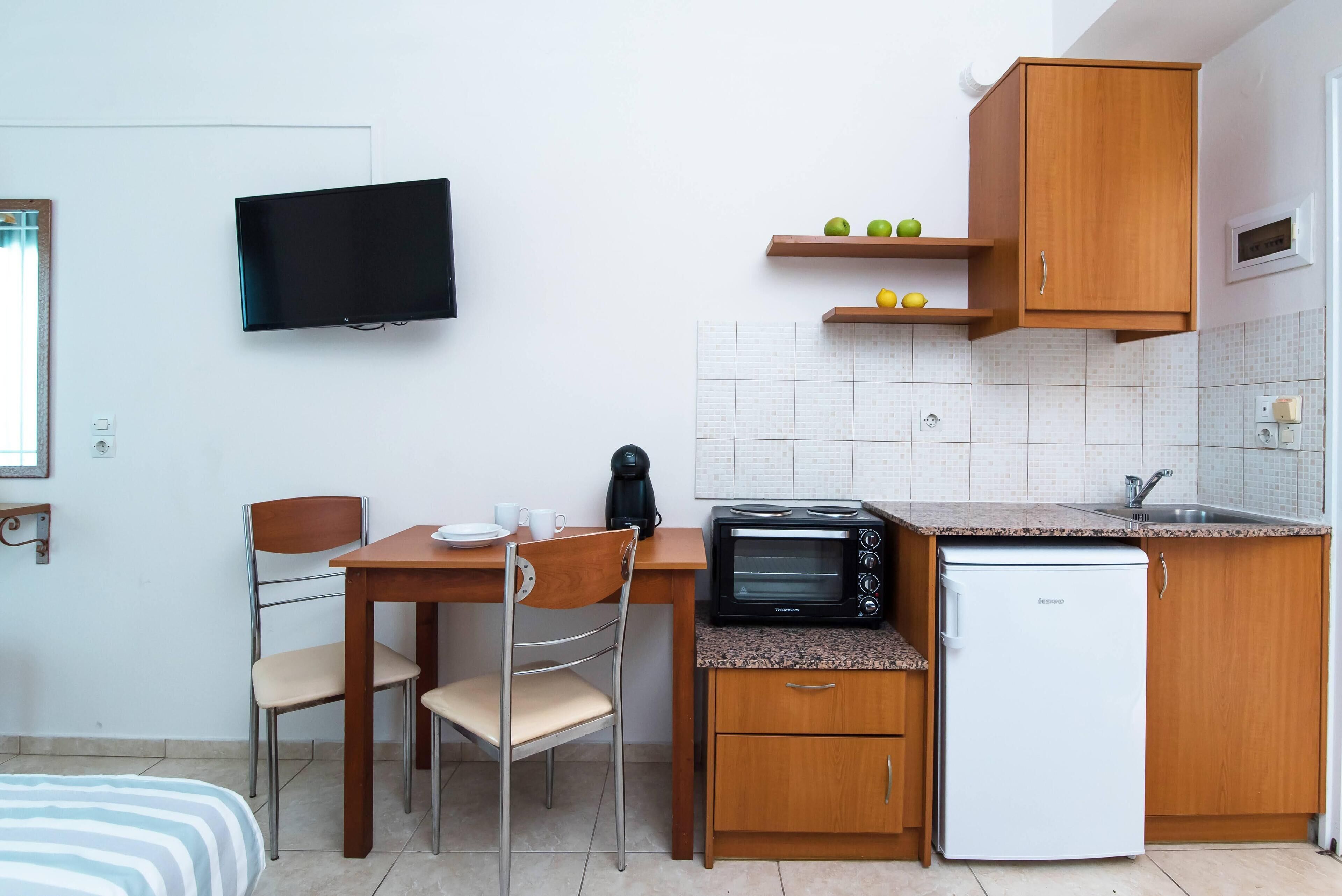 A compact kitchenette with a microwave, toaster, and refrigerator, a wooden dining table with chairs, a television mounted on the wall, and a small shelf with fruits.