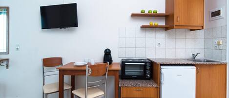 A compact kitchenette with a microwave, toaster, and refrigerator, a wooden dining table with chairs, a television mounted on the wall, and a small shelf with fruits.