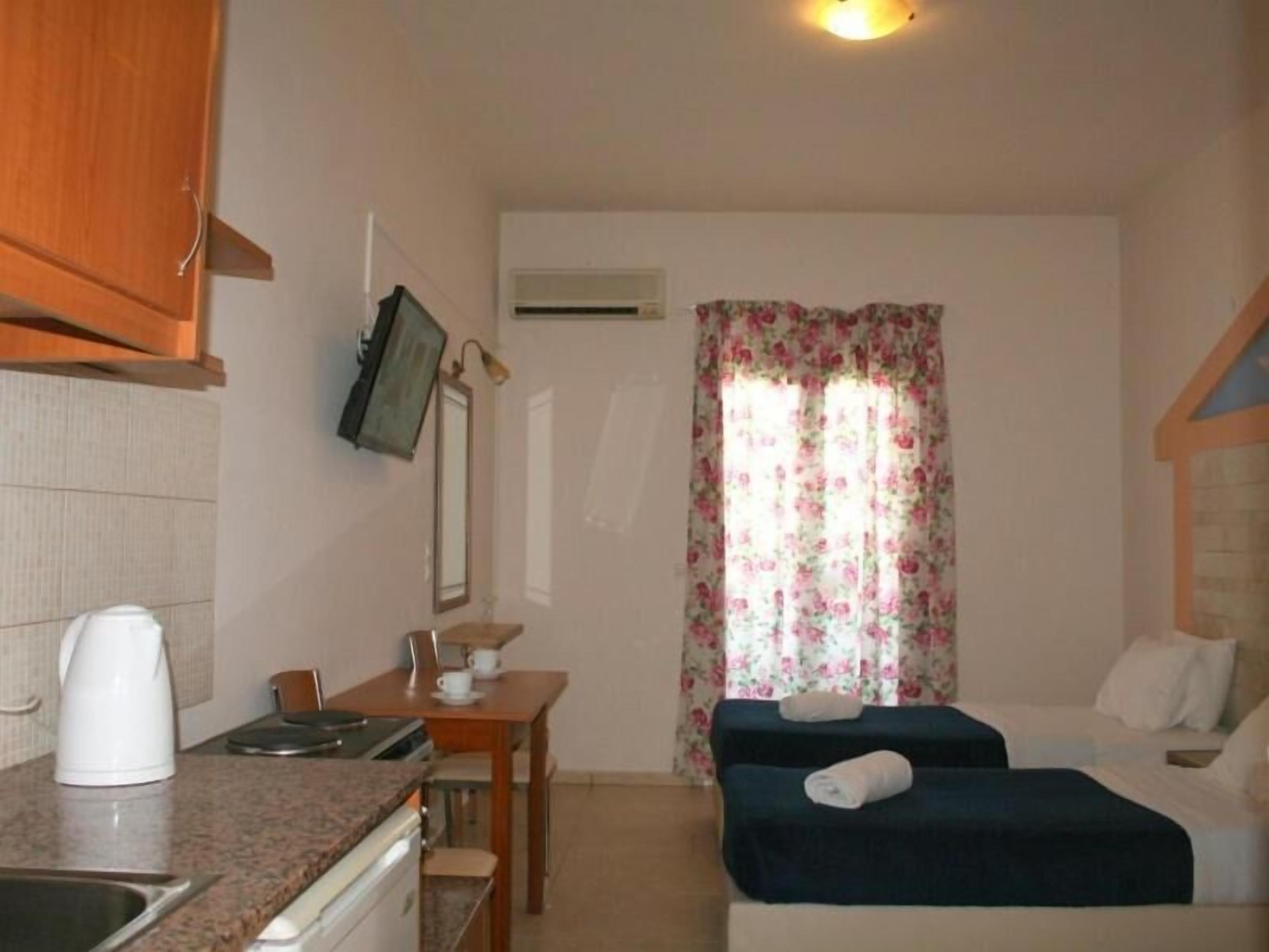 Une pièce compacte comprenant un coin cuisine, un lit, une petite table avec une chaise et une fenêtre agrémentée de rideaux à motifs floraux.