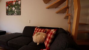 Un salón con un sofá gris oscuro, una manta roja y blanca a cuadros y un almohadón blanco mullido. Un cuadro de flores cuelga en la pared.