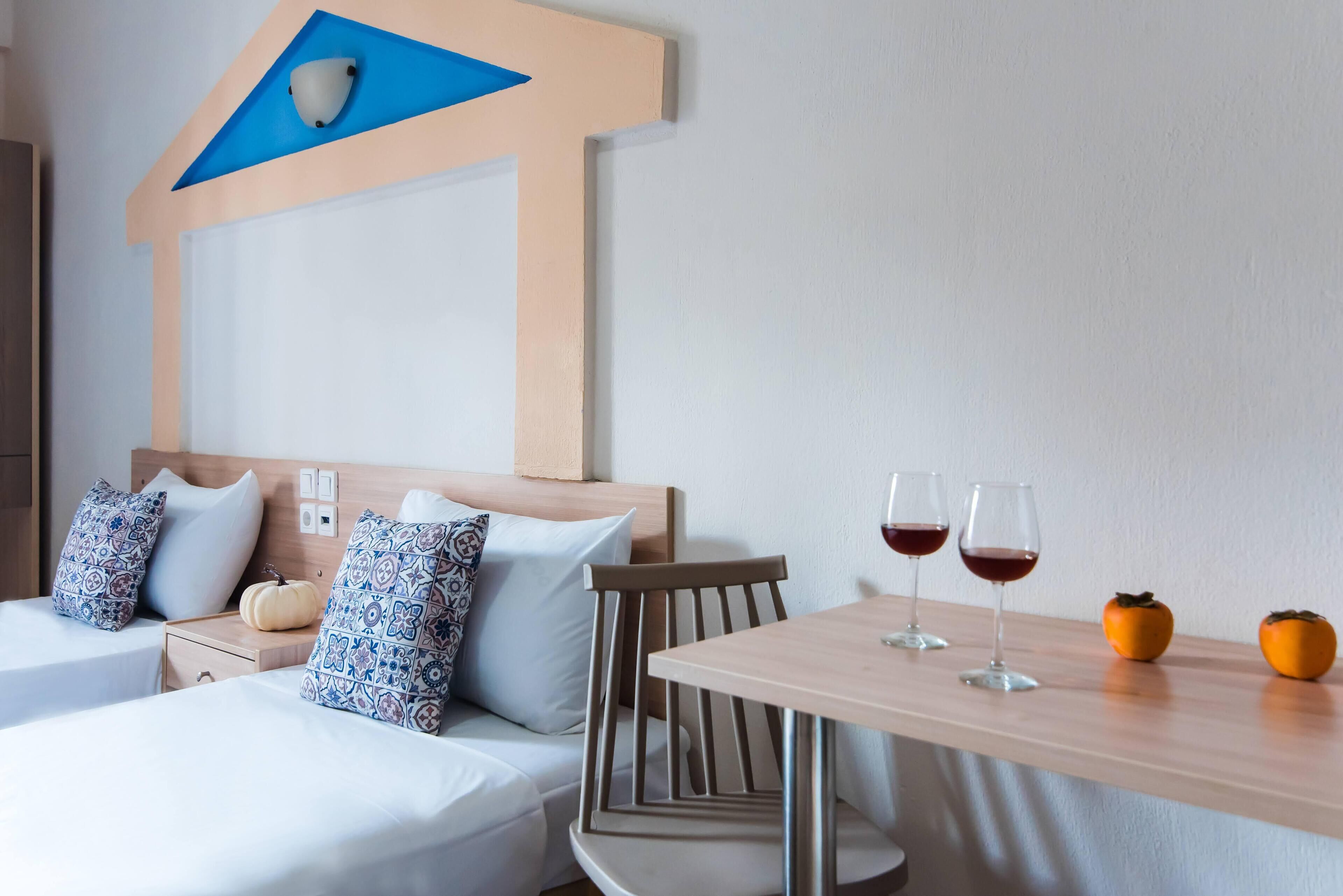 A hotel room with two beds, a wooden headboard, a nightstand, a table with two wine glasses, and two small pumpkins.