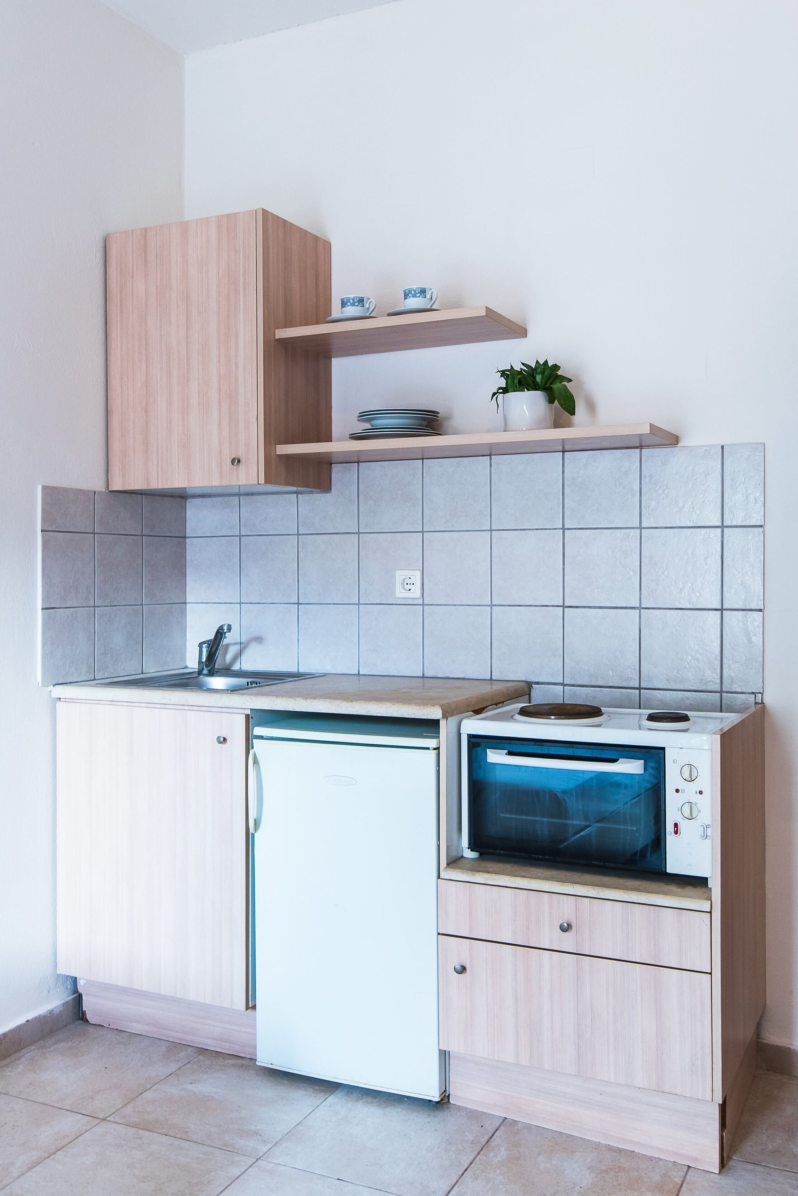 A compact kitchen with a built-in oven, a small sink, and a microwave. There is a wooden cabinet above the sink and a shelf with dishes and a plant.