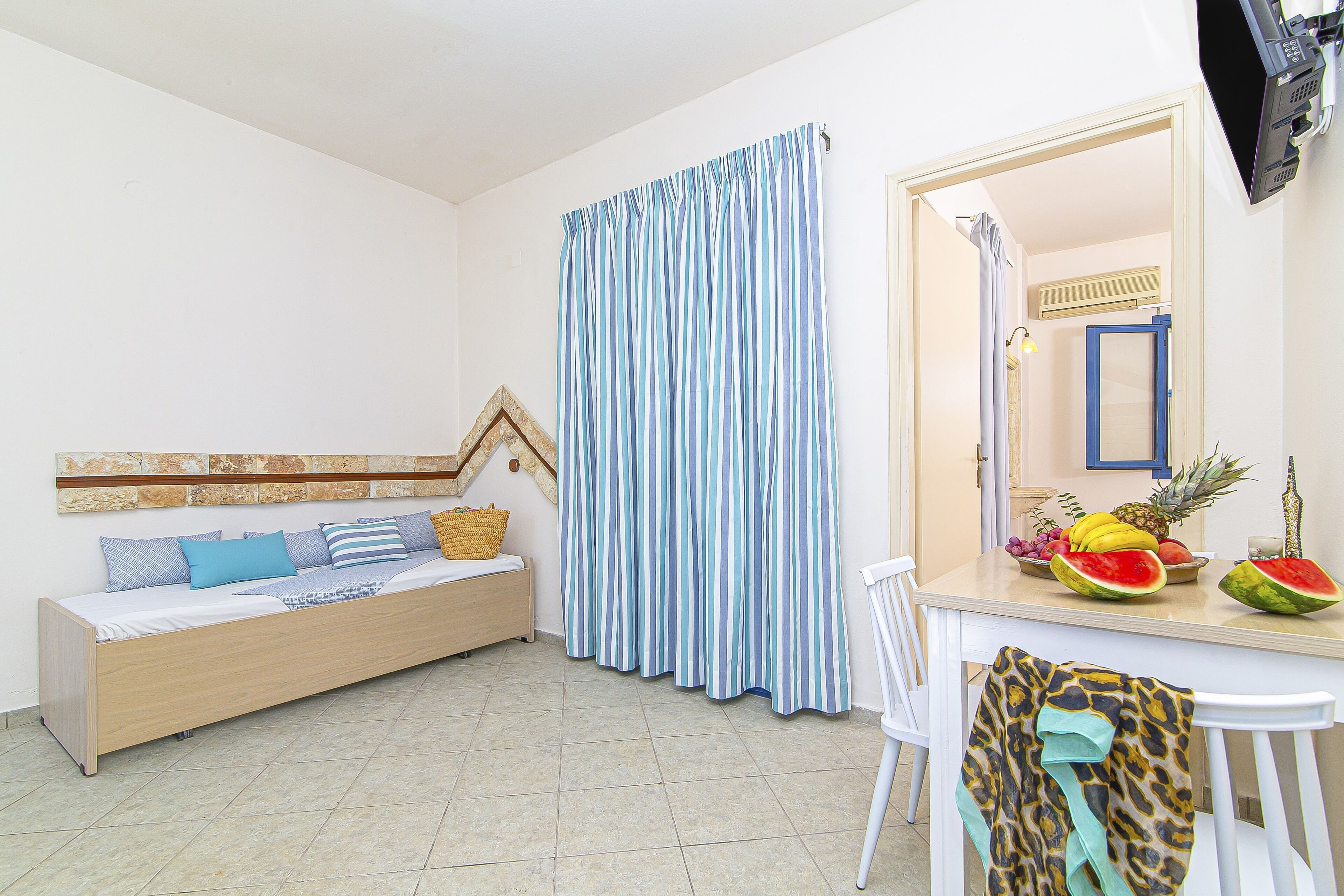 A room with a bed, a dining table with fruits, a white chair, and a striped curtain.