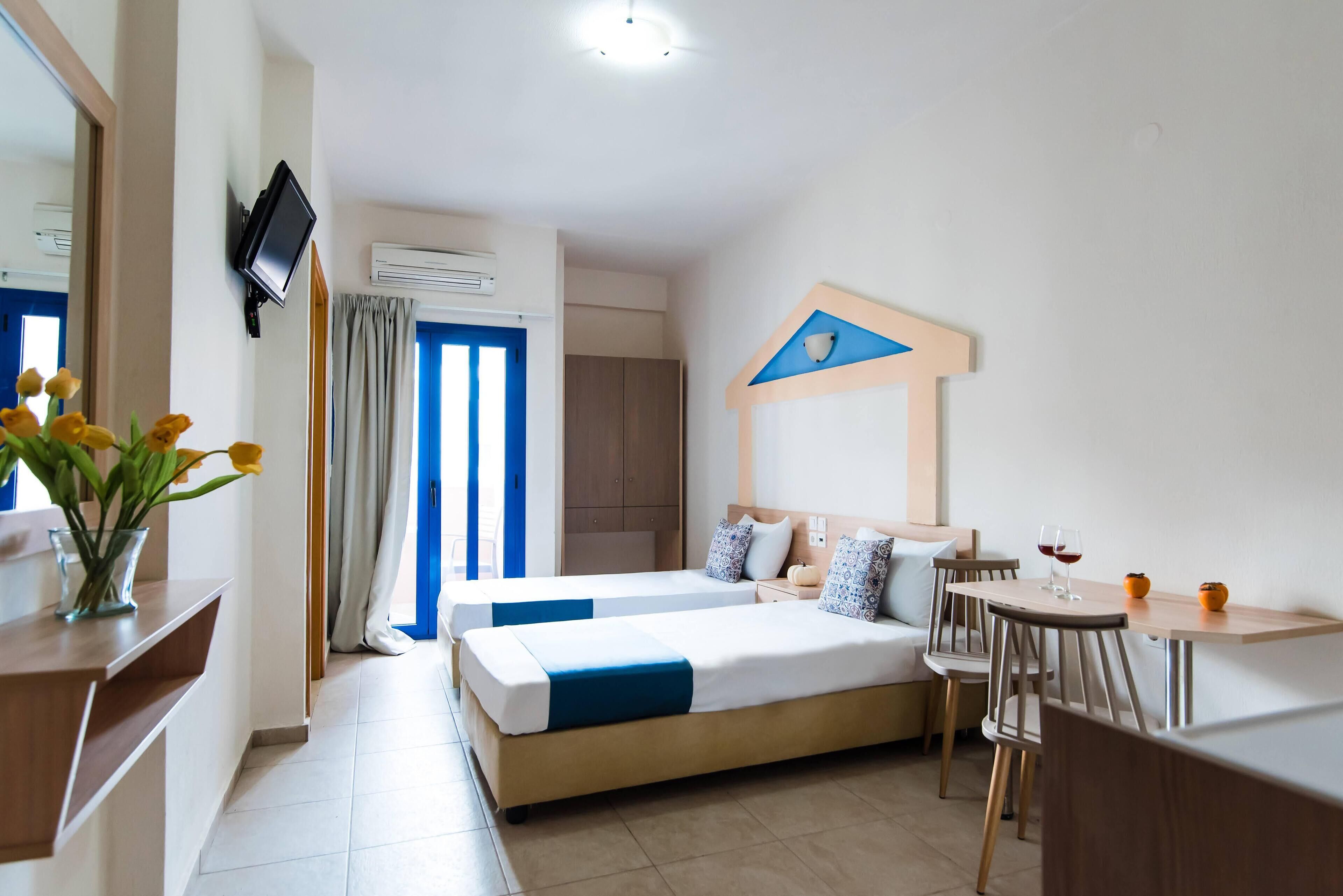 A hotel room with two beds, a TV mounted on the wall, a desk with a chair, and a table with two glasses of red wine.