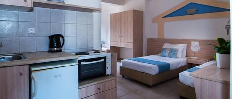 Habitación de hotel compacta con kitchenette, cama con almohada azul y blanca a rayas, y una mesita de noche con lámpara.