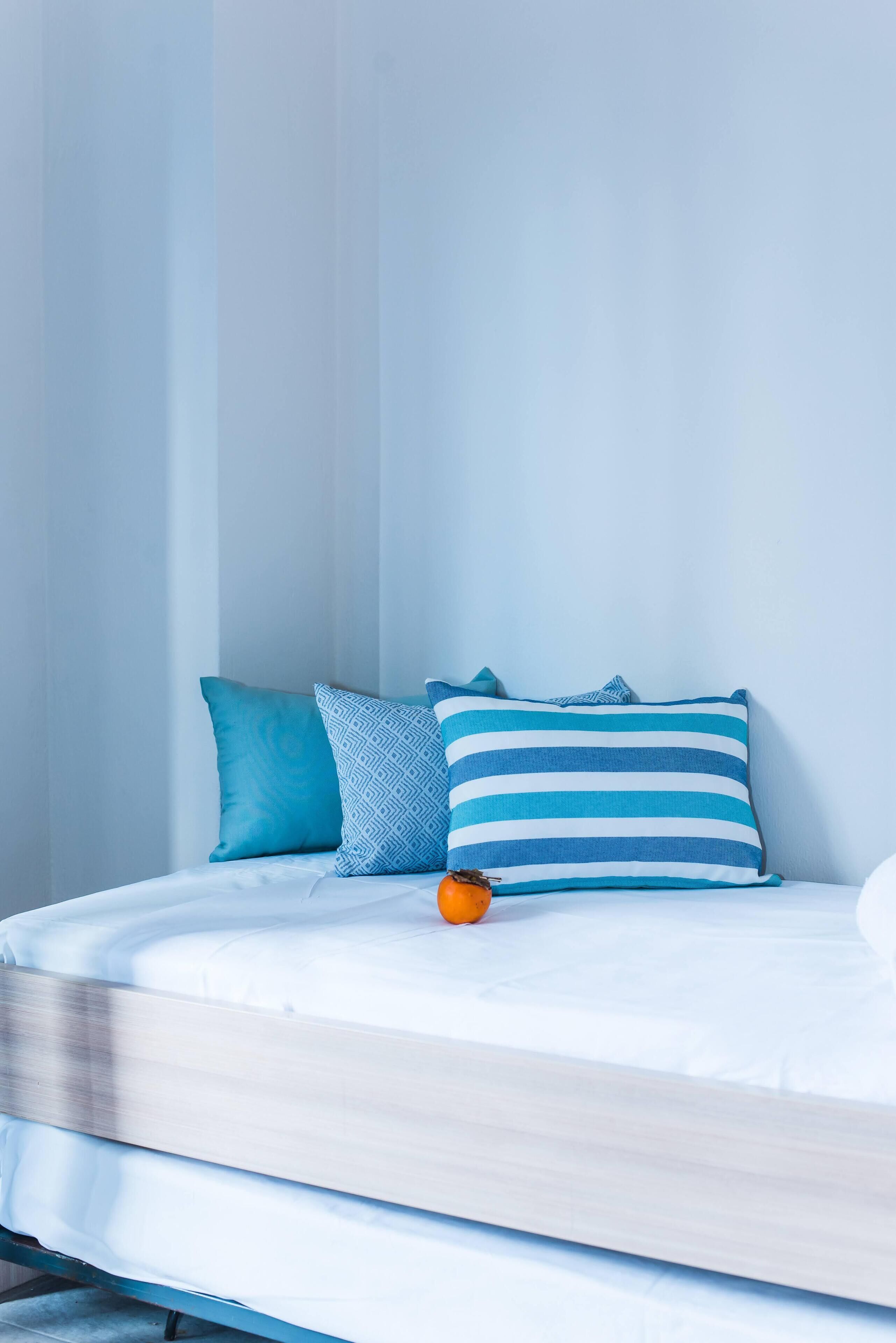 A bed with white bedding, two pillows, one with a blue geometric pattern and the other with blue stripes, and an orange object on the bed.