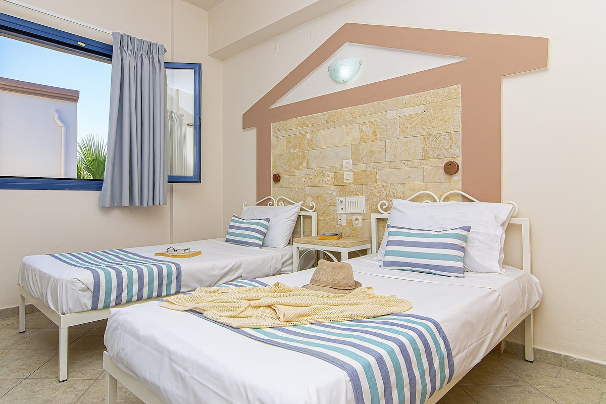 A hotel room with two beds, a window with curtains, a stone wall, and a ceiling light.