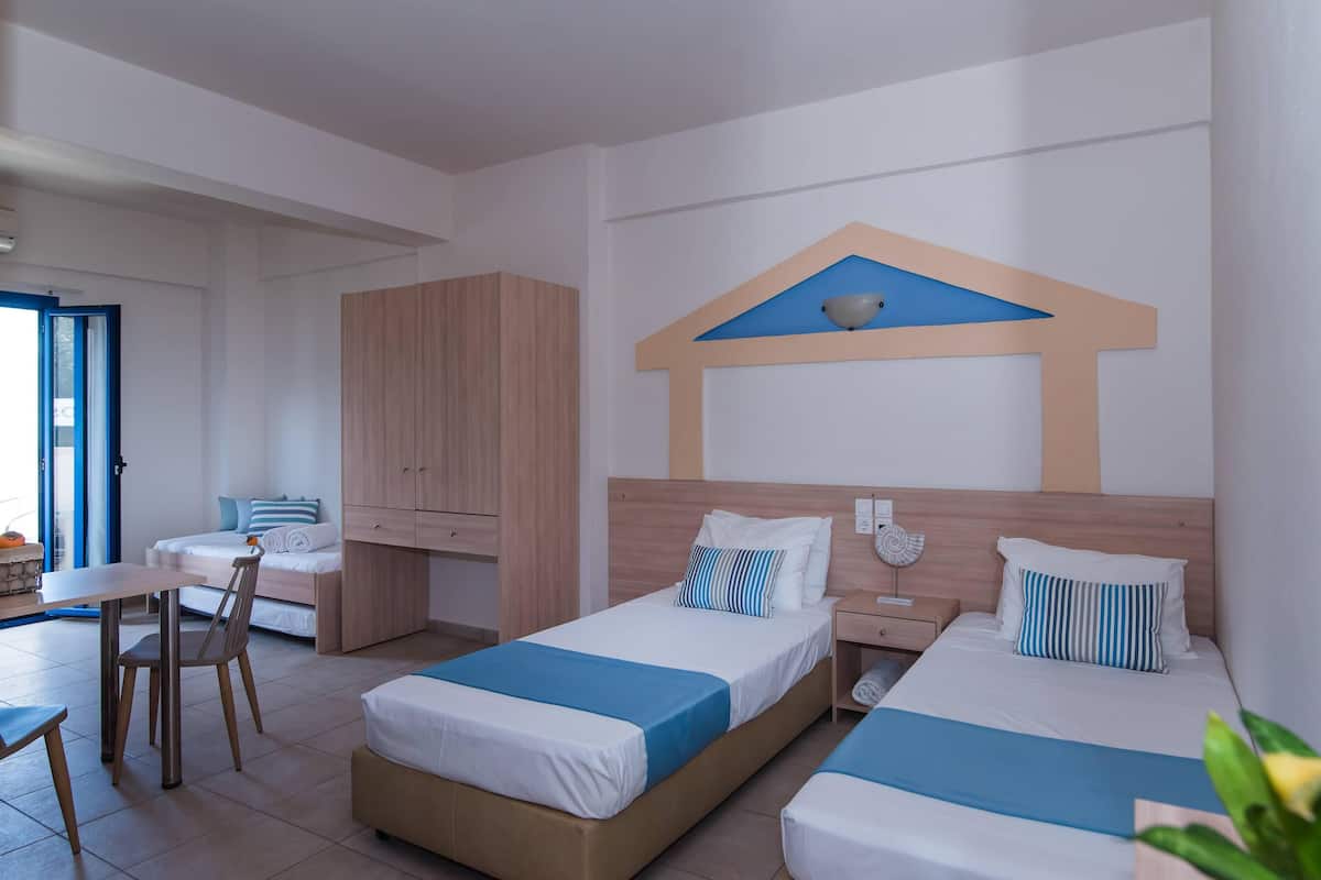 A hotel room with two beds, a wooden wardrobe, a dining table with chairs, and a view of the sea through a large window.