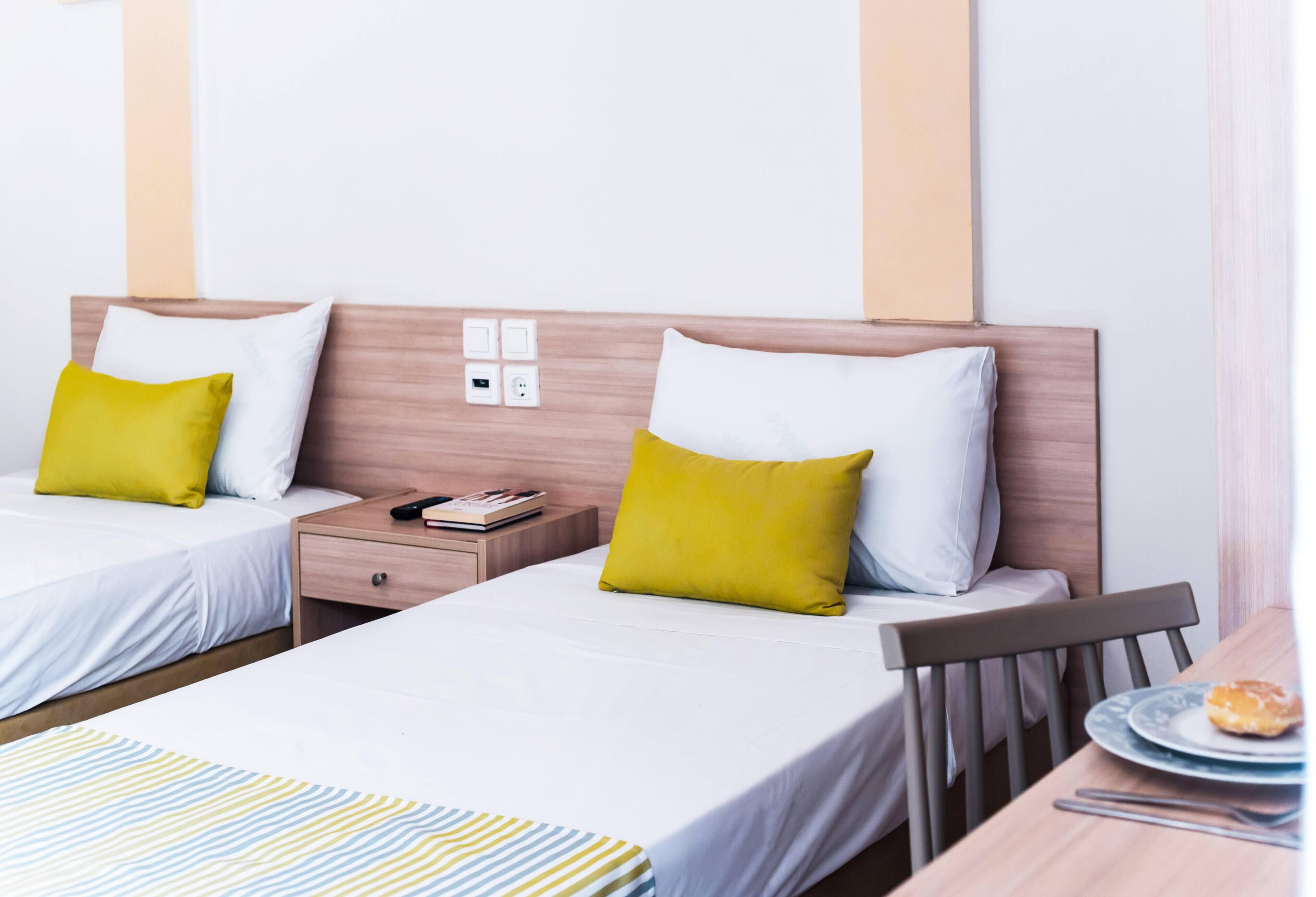 A hotel room with two beds, each with a white mattress and a pillow. There is a wooden headboard, a bedside table with a book and a remote control, and a small table with a plate and a pastry.