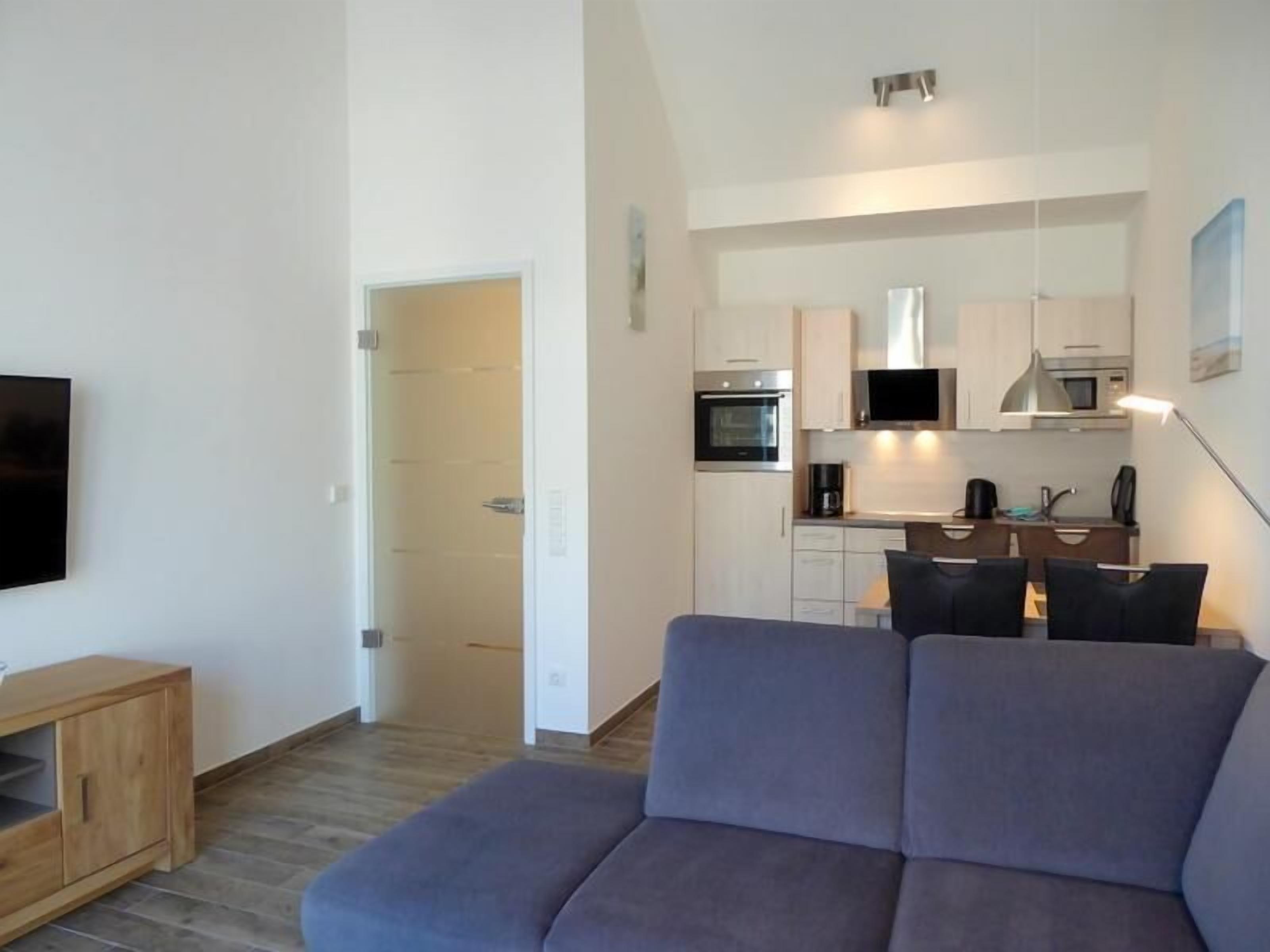 A modern living room with a grey sofa, a wooden cabinet, and a kitchen area with white cabinets, a microwave, and a TV.