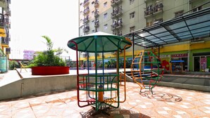 An outdoor playground with swings and a covered seating area, surrounded by a tiled plaza and residential buildings. - The Suites Metro Apartment by NFO Property (Bandung)