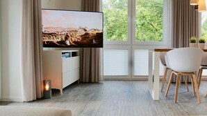 Moderni olohuone, jossa on seinälle asennettu taulutelevisio valkoisen lipaston päällä, ruokailutila puupöydän ja -tuolien kera sekä suuret ikkunat, joista avautuu näkymä vehreään maisemaan.