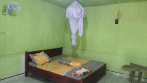 Quarto com cama, mosquiteiro suspenso, mesinha e ventoinha fixada na parede.