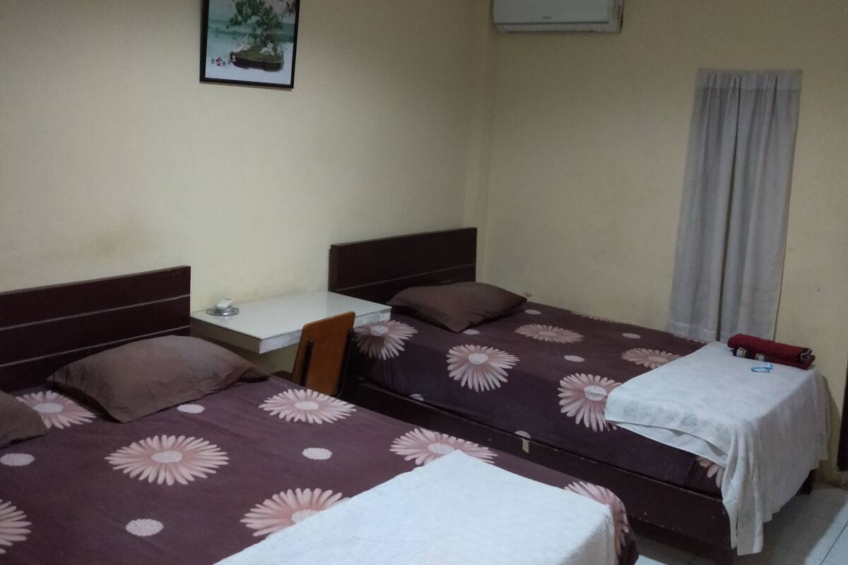 A hotel room with two beds, a desk, a chair, an air conditioning unit, and a window with curtains.