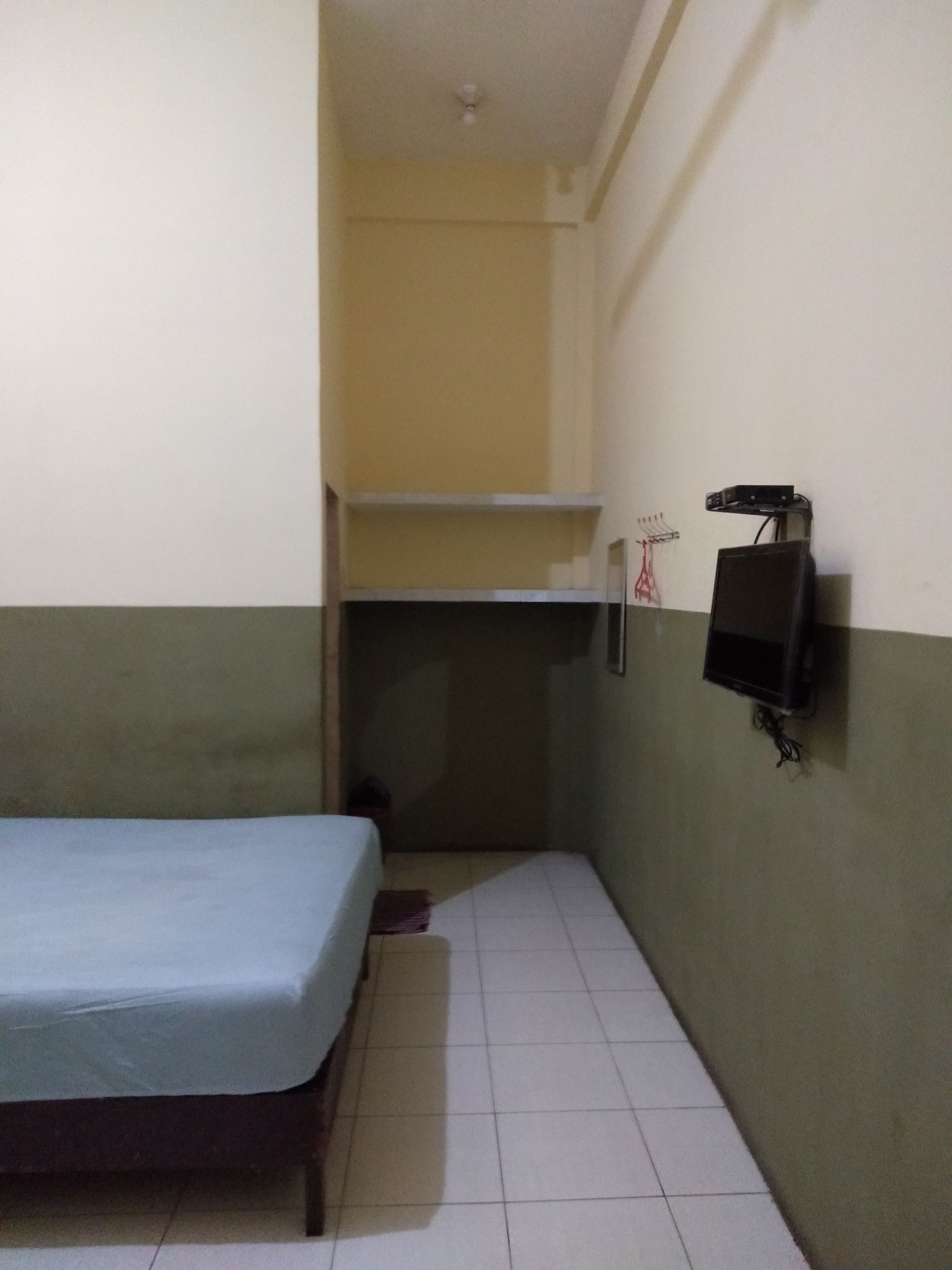 A small, narrow room with a single bed, a shelf, and a wall-mounted TV.
